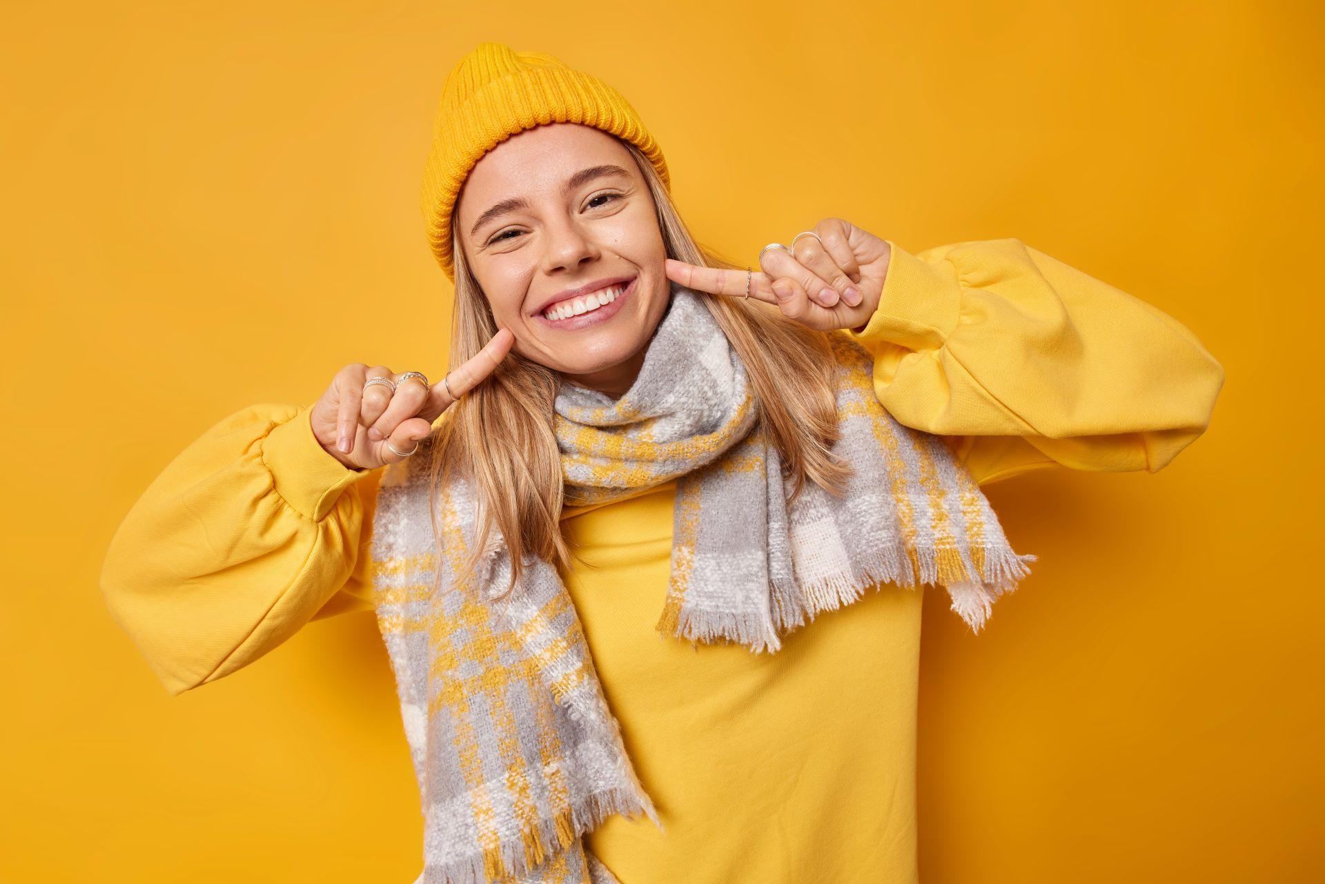 Eine junge Frau lächelt fröhlich in die Kamera und zeigt mit beiden Zeigefingern auf ihre Zähne. Sie trägt eine gelbe Mütze, einen gelben Pullover und einen grauen Schal mit gelbem Muster. Der Hintergrund ist komplett gelb, passend zu ihrer Kleidung.